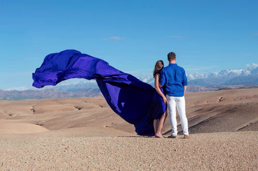Séance photo romantique en robe volante pour couple dans le désert d'Agafay près de Marrakech, au Maroc. La femme porte une robe bleu royal spectaculaire avec une traîne imposante qui flotte au vent, tandis qu'elle pose avec son partenaire, avec en toile de fond les majestueuses montagnes de l'Atlas enneigées. Une curation de Marrakech Lux Events.