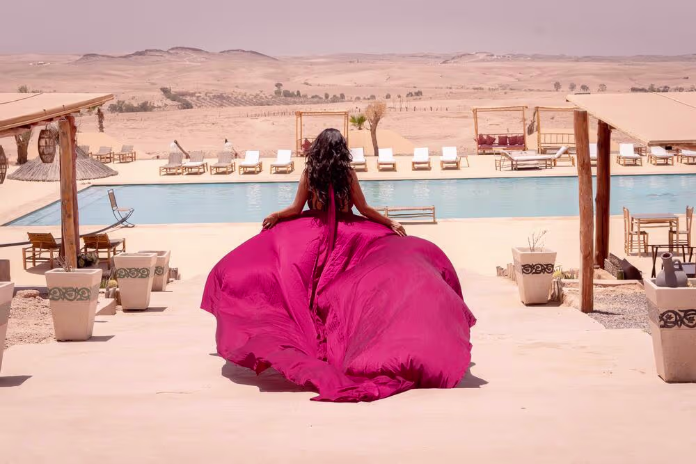 Vue arrière dramatique d'une séance photo en robe volante dans un resort du désert d'Agafay, au Maroc. Le mannequin est assis au bord de la piscine bleue, sa robe volumineuse magenta s'étalant sur la terrasse, avec vue sur les dunes et le salon extérieur. Une curation de Marrakech Lux Events.