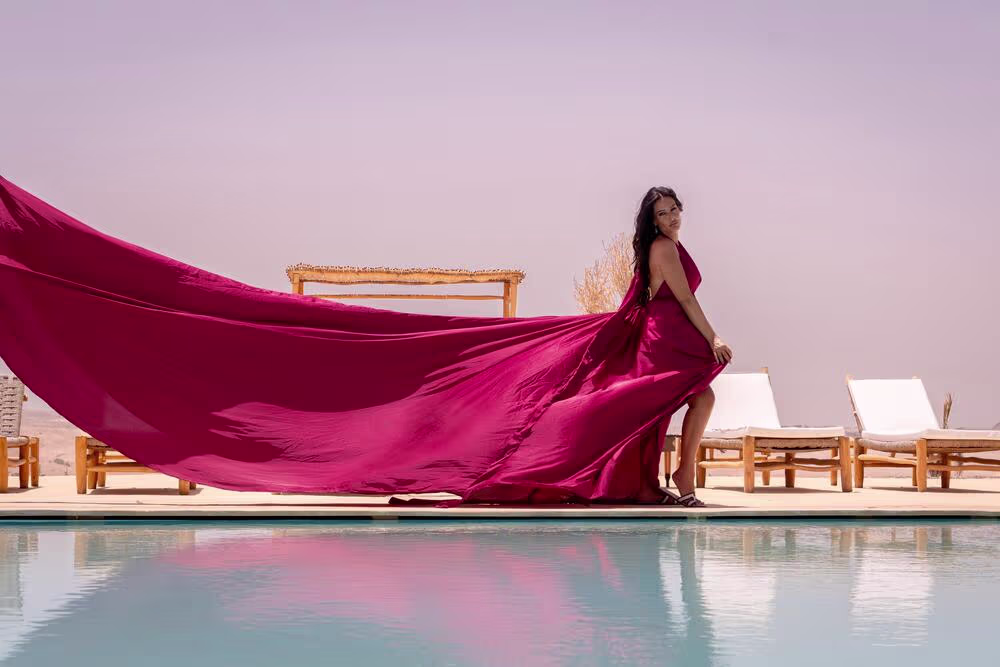 Séance photo dynamique en robe volante d'un magenta éclatant au bord de la piscine d'un resort de luxe dans le désert d'Agafay, au Maroc. Le tissu fuchsia flotte dramatiquement sous l'effet du vent tandis que le mannequin pose devant des transats en bois blanc. Une réalisation de Marrakech Lux Events.