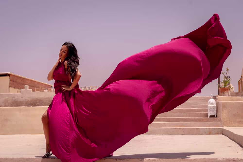 Séance photo chic en robe volante sur un toit traditionnel marocain, mettant en valeur une robe spectaculaire de couleur magenta. Le tissu vibrant se soulève élégamment sous le vent tandis que le mannequin pose sur des escaliers en pierre, à côté d'une lanterne marocaine blanche, sous un ciel dégagé. Une création de Marrakech Lux Events.