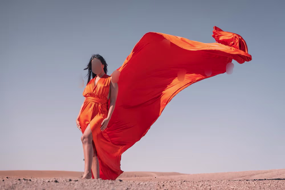 Shooting photo éditorial spectaculaire en robe volante dans le désert d'Agafay, au Maroc. Le mannequin pose avec assurance dans une magnifique robe orange flamboyant qui virevolte dans le vent, face à un ciel bleu dégagé et aux paysages minéraux du désert. Imaginé par Marrakech Lux Events.