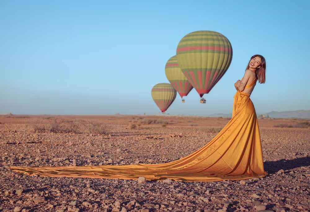 Séance photo époustouflante en robe volante dans le désert rocailleux d'Agafay, au Maroc. Une mannequin pose dans une robe jaune spectaculaire à la traîne virevoltante, sur un terrain accidenté, tandis que trois montgolfières colorées flottent dans le ciel bleu azur. Une création signée Marrakech Lux Events.