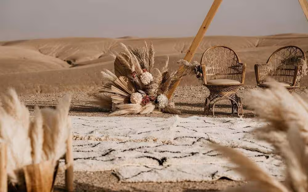 Pique-nique luxueux et bohème dans le désert d'Agafay, Maroc. Un couple installé dans deux célèbres fauteuils paon en rotin, posés sur un tapis marocain noir et blanc à motifs, avec au centre un arrangement de fleurs séchées mêlant pampa et palmes, imaginé par Marrakech Lux Events pour un dîner romantique en plein désert.