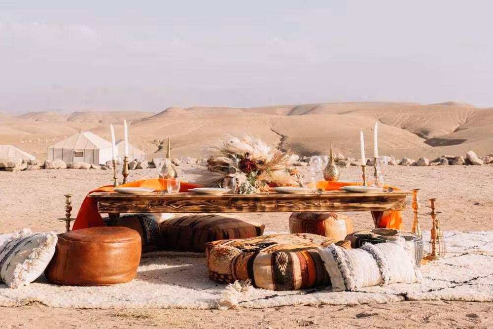 Pique-nique luxueux en journée dans le désert rocailleux d'Agafay, Maroc. Une table basse en bois rustique dressée avec assiettes et verres, entourée de poufs marocains en cuir marron et en tresses. Au centre, une composition de fleurs séchées, des bougeoirs en laiton et, en fond, des tentes blanches de glamping qui se détachent sur le paysage désertique. Un concept signé Marrakech Lux Events.