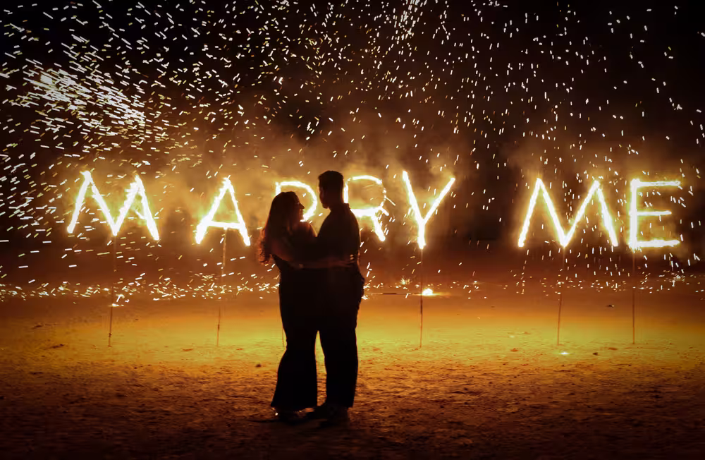 Demande en mariage nocturne spectaculaire dans le désert de Marrakech avec un couple en silhouette devant une enseigne enflammée « MARRY ME » et une pluie d'étincelles d'artifices dorées, conçue par Marrakech Lux Events.