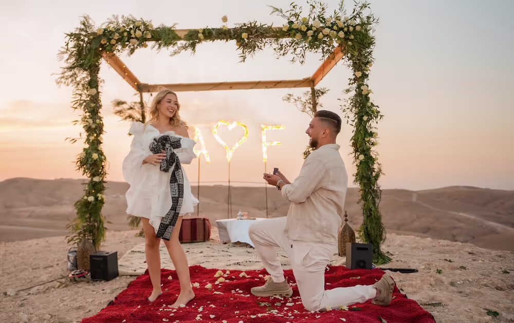 Demande en mariage luxueuse au coucher du soleil dans le désert d'Agafay avec un homme à genoux sur un tapis rouge sous une arche florale romantique, avec des lettres lumineuses « LOVE » et un service de champagne en arrière-plan, créée par Marrakech Lux Events.